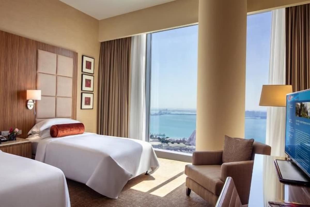 A hotel room with two beds, a desk, a chair, a television, and a large window with a view of the sea.