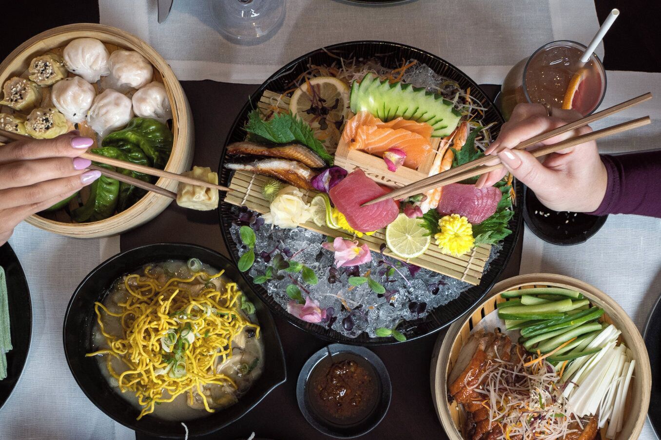 A table with various dishes including dumplings, sashimi, noodles, and a bowl of soup.
