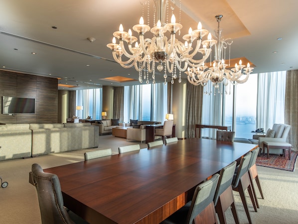 A spacious hotel room with a large dining table, comfortable seating, a modern kitchen area, and large windows offering a city view.