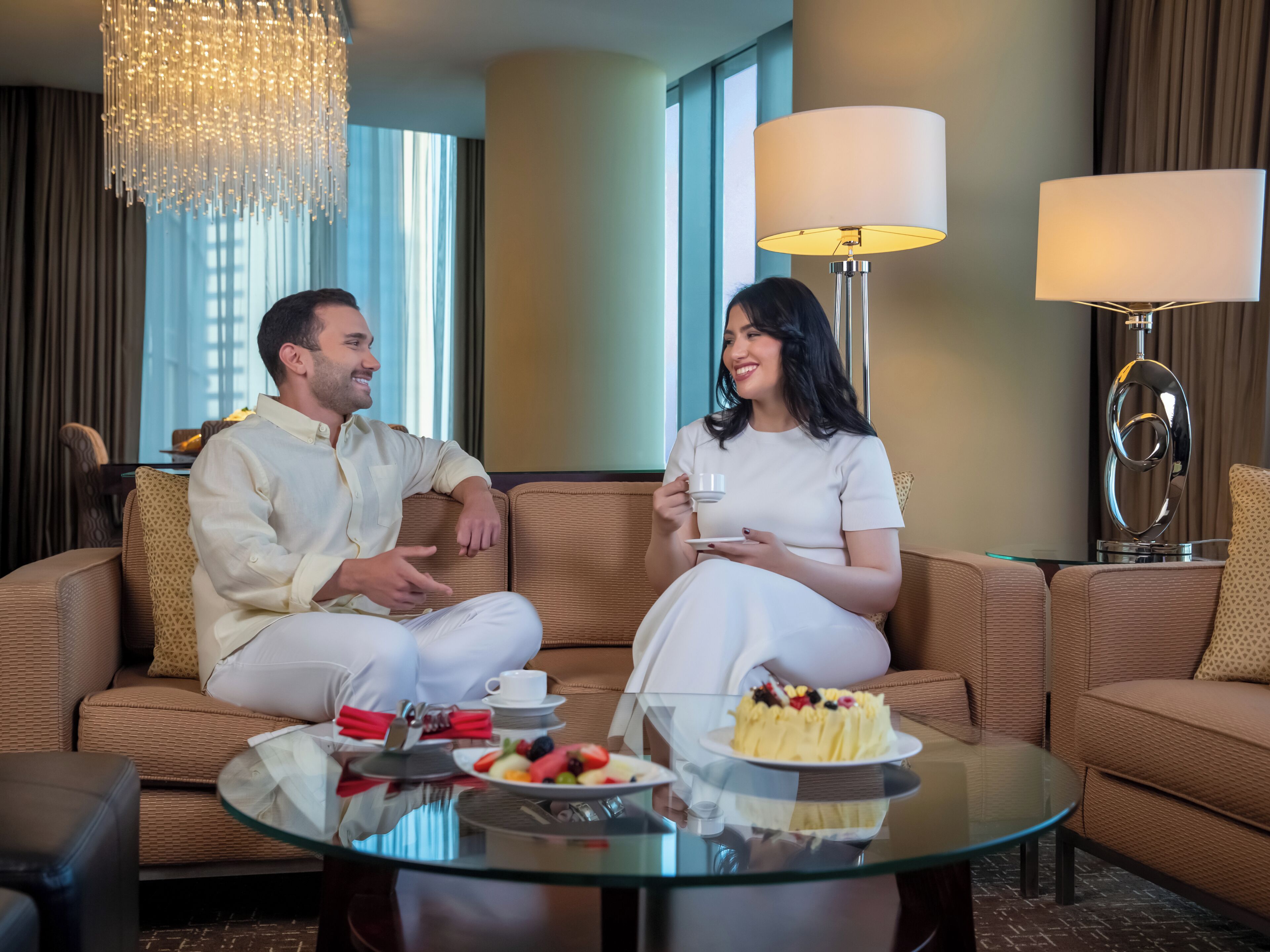 Una pareja sentada en un sofá marrón en una habitación bien iluminada, con una mesa de centro de vidrio, un pastel y una bandeja de frutas.