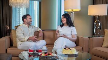 Una pareja sentada en un sofá marrón en una habitación bien iluminada, con una mesa de centro de vidrio, un pastel y una bandeja de frutas.