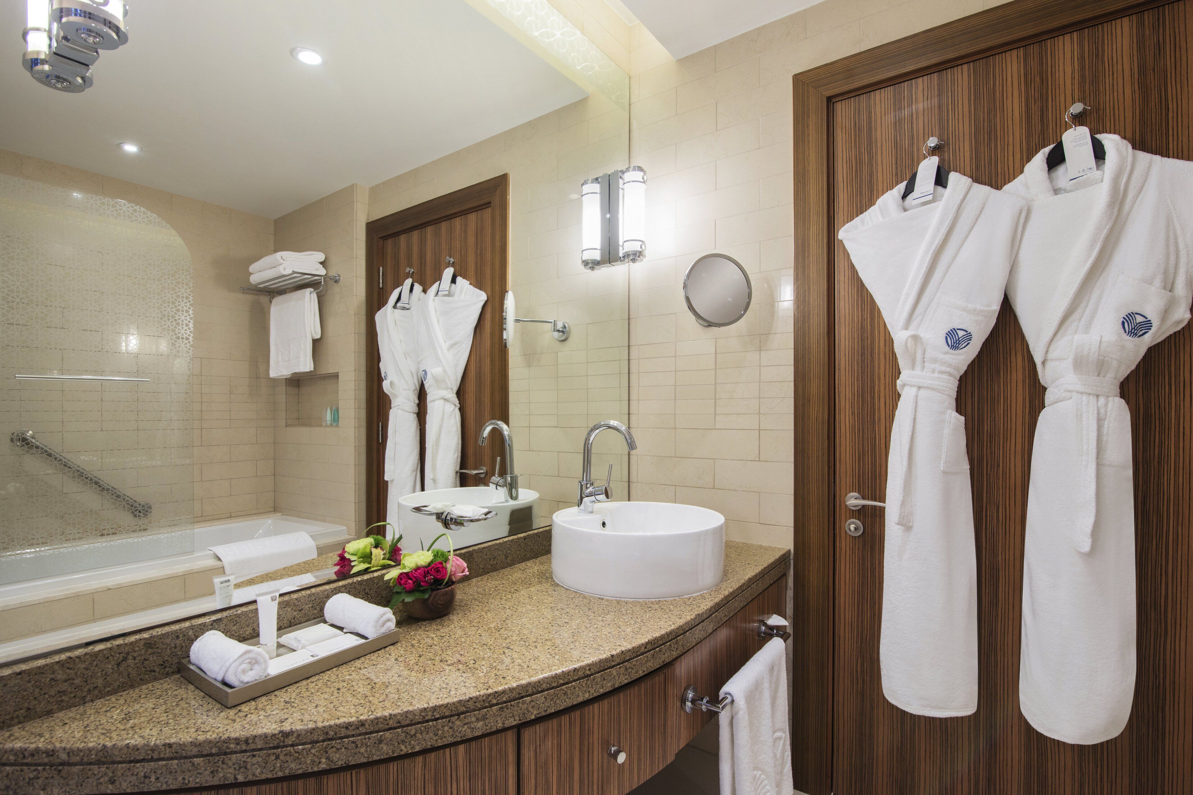 A modern bathroom with a bathtub, sink, and mirror. There are towels and robes hanging on the wall.