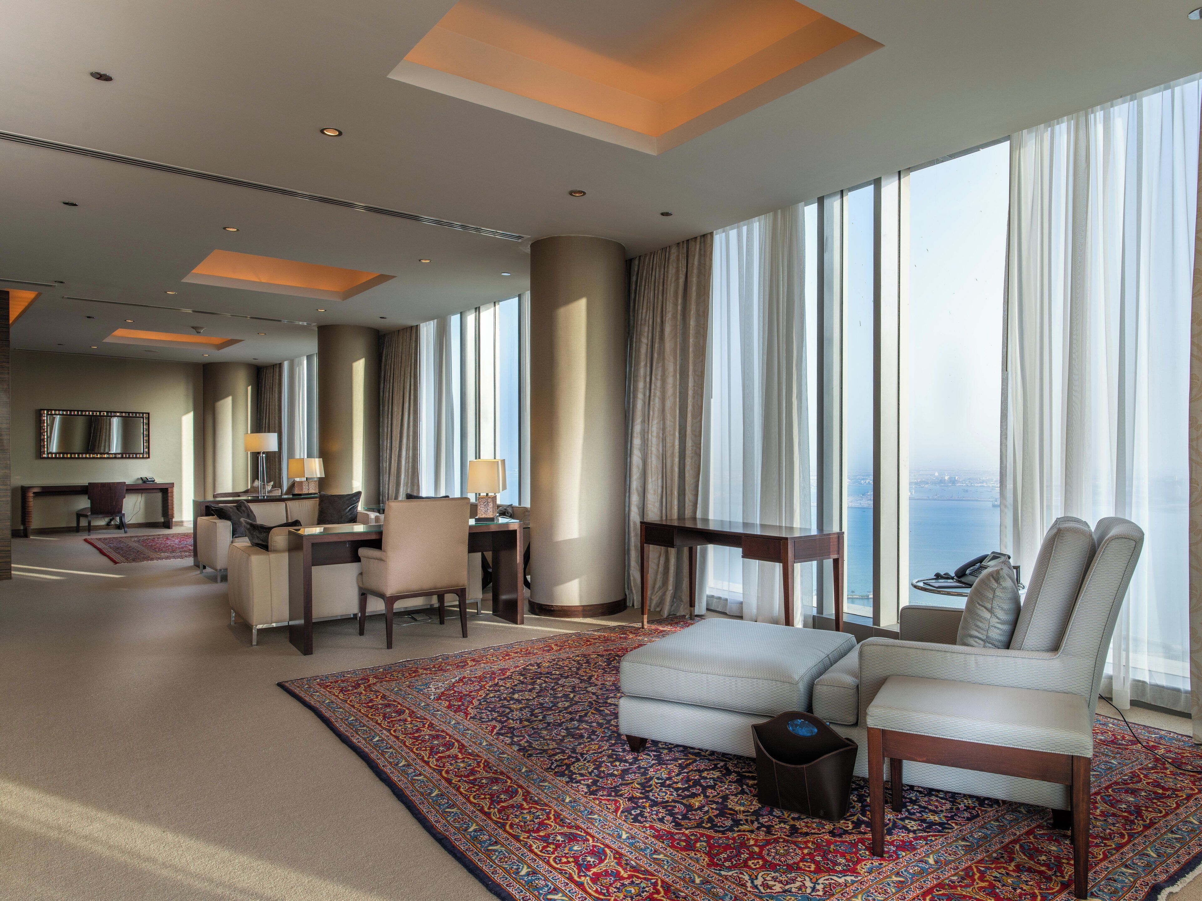 A spacious hotel room with a large window, a sitting area with a sofa and armchairs, a desk with a chair, and a large rug.