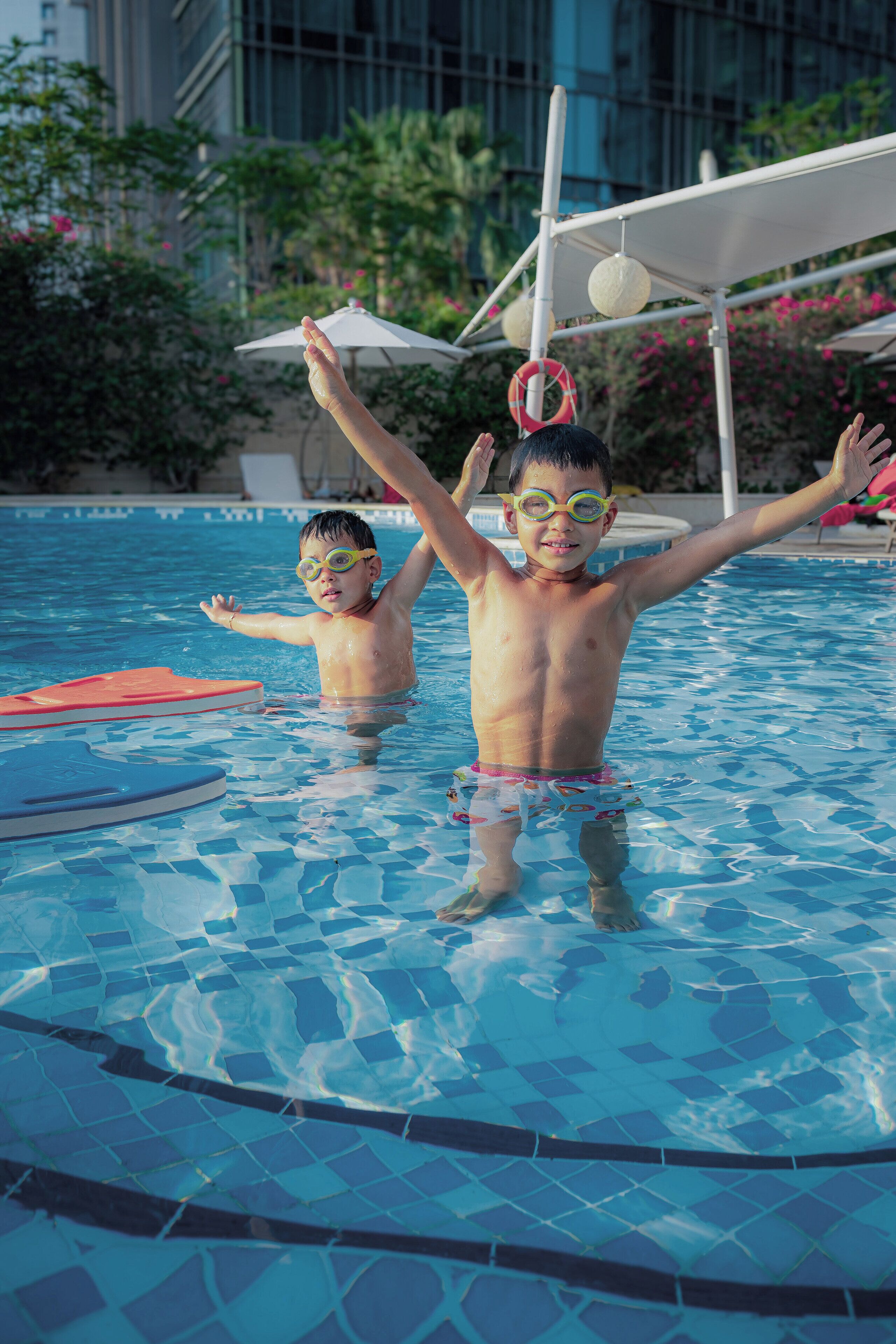 Duas crianças em uma piscina com piso de azulejos e uma estrutura de guarda-sol à beira da piscina.