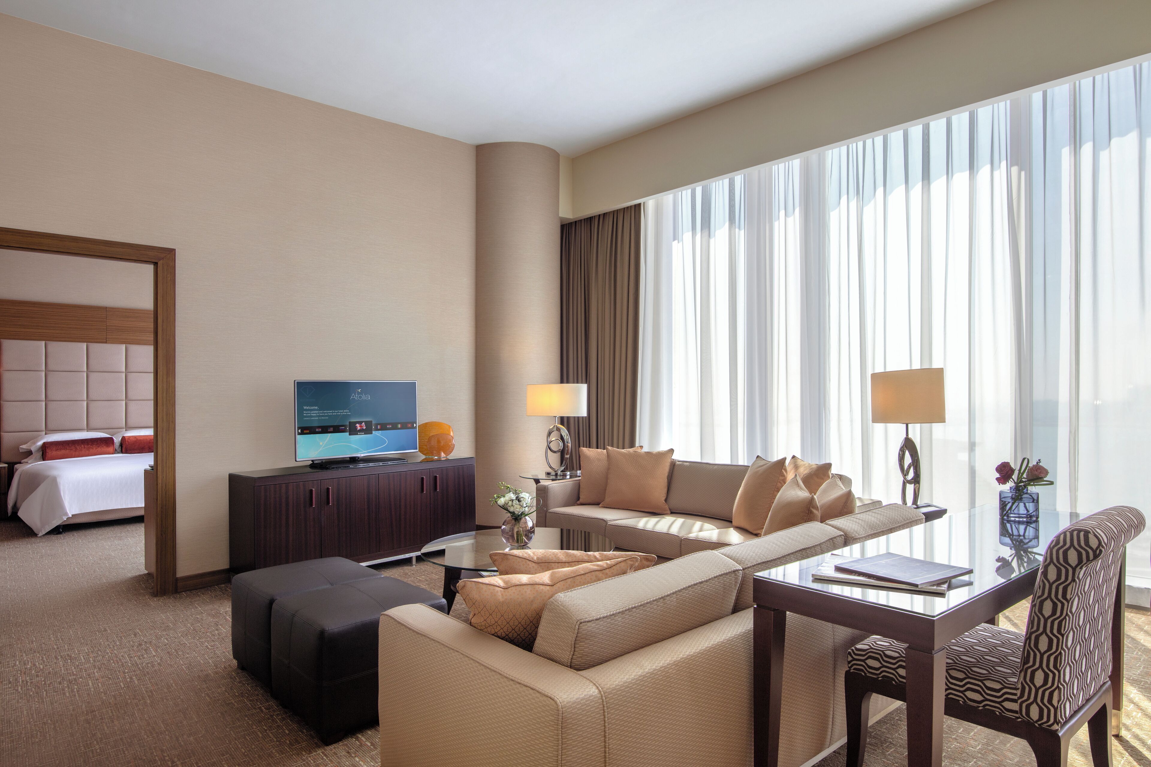 Habitación de hotel con una cama grande, un sofá, una mesa de centro, un televisor y vistas al exterior a través de amplios ventanales.