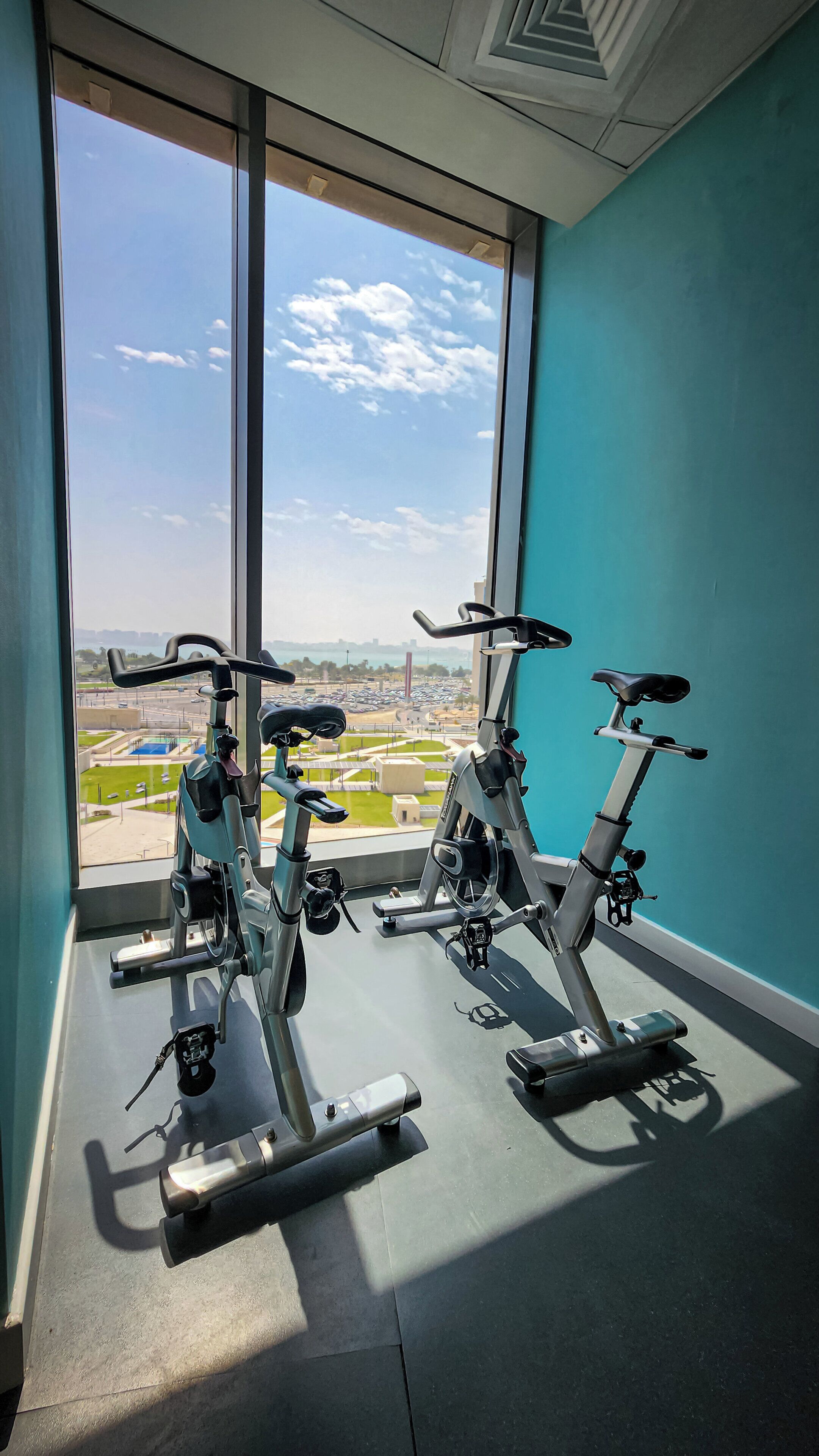 A gym with two stationary bikes, large windows, and a view of a parking lot and buildings.