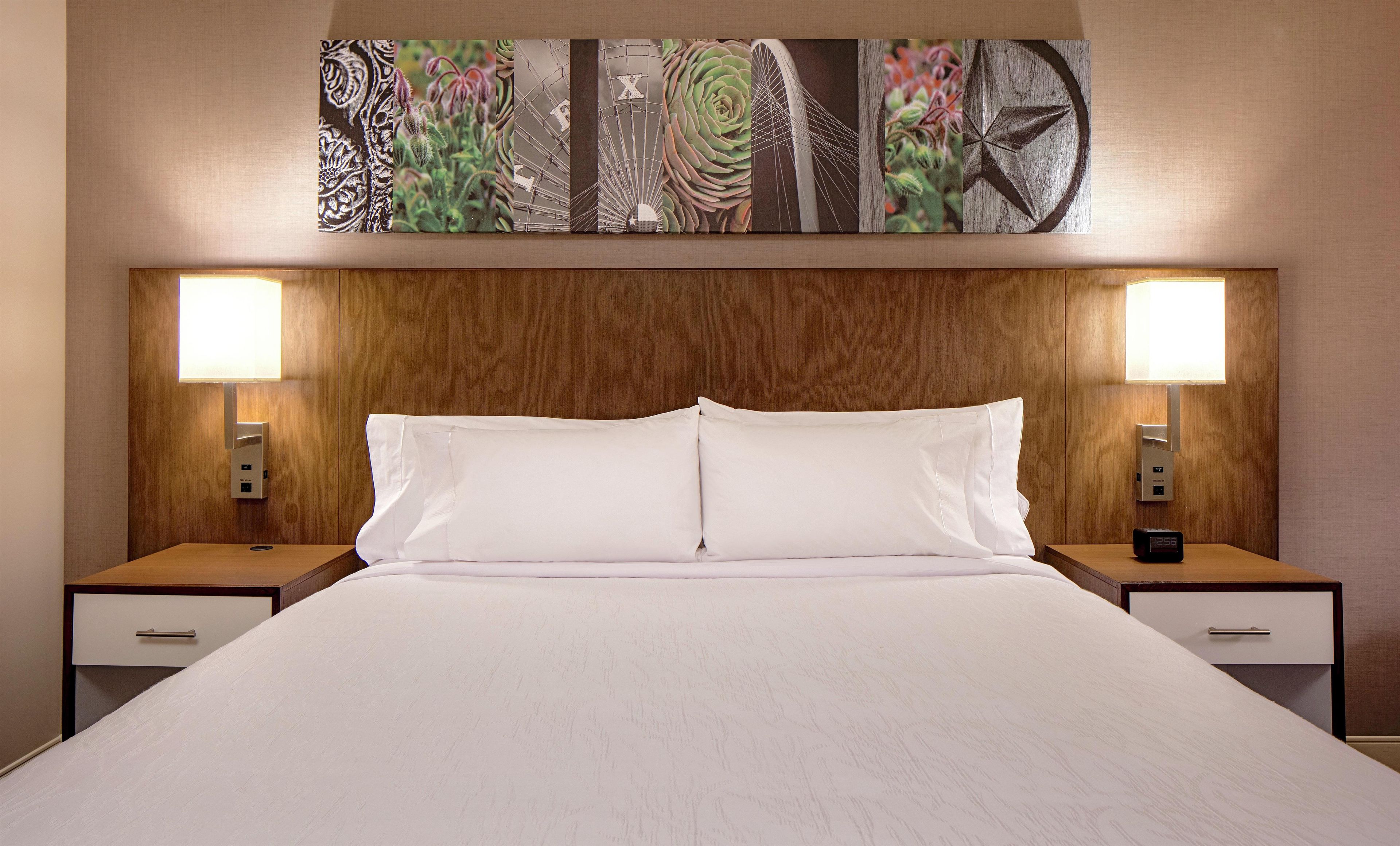 A hotel room with a large bed, two bedside tables, a nightstand with a clock, and a wall-mounted artwork above the bed.