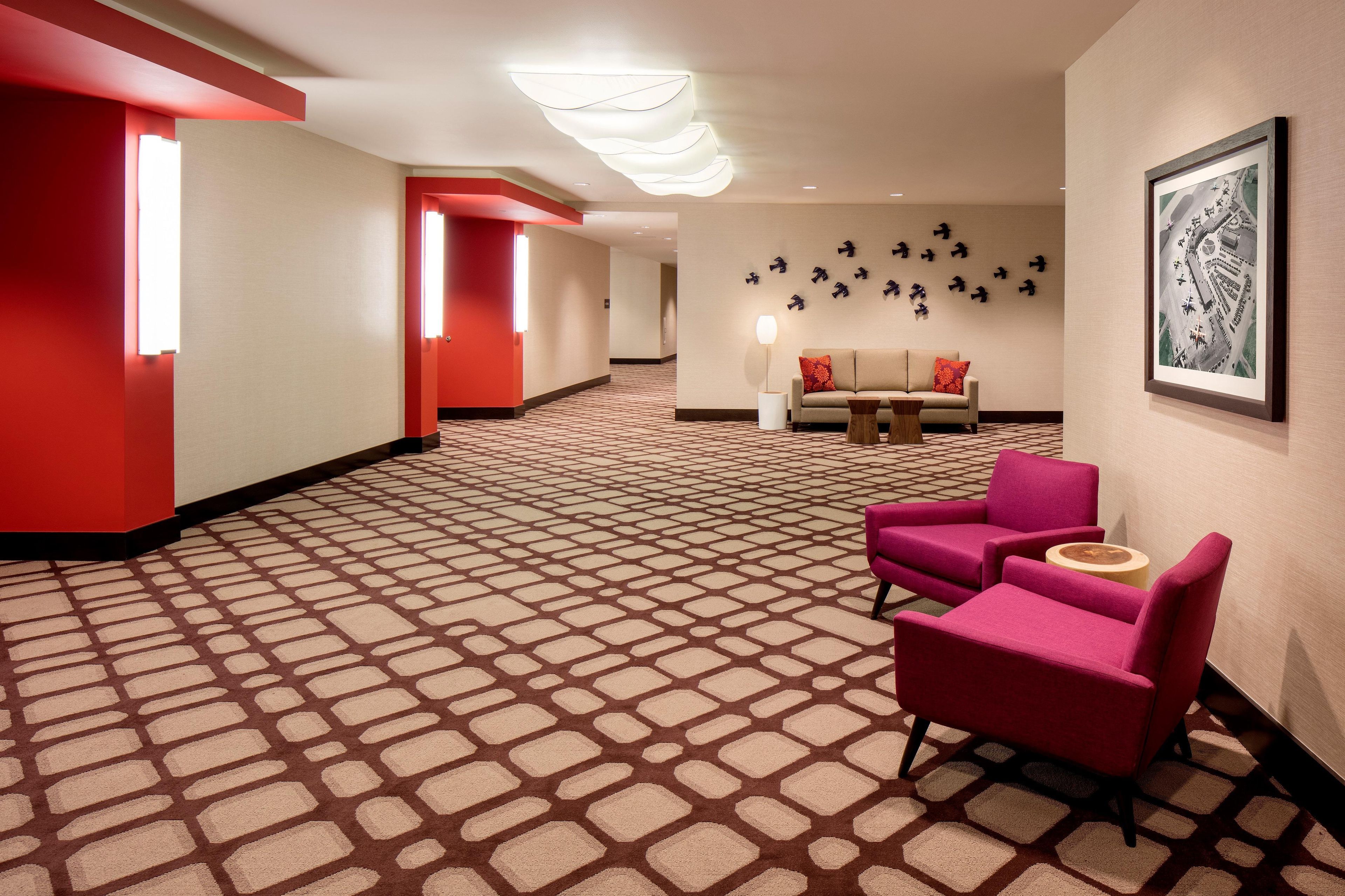 A hotel lobby with a patterned carpet, red walls, a sofa, two armchairs, and a wall-mounted art piece.