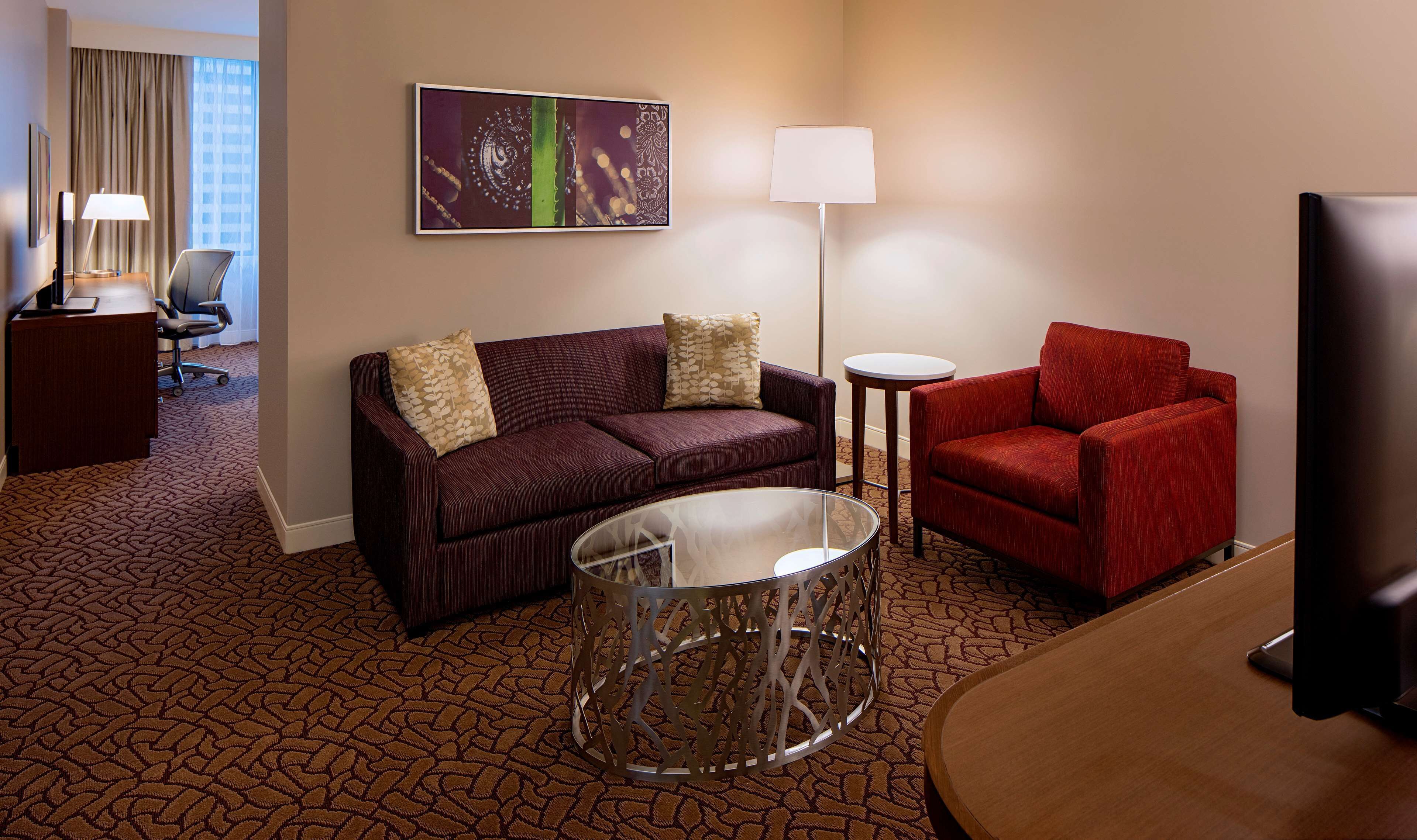 A hotel room with a dark brown patterned carpet, a dark brown sofa with patterned cushions, a red armchair, a small round table, a glass coffee table, a flat-screen TV, a desk with a lamp, and a framed abstract artwork on the wall.