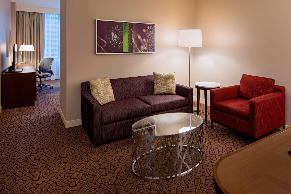 A hotel room with a dark brown patterned carpet, a dark brown sofa with patterned cushions, a red armchair, a small round table, a glass coffee table, a flat-screen TV, a desk with a lamp, and a framed abstract artwork on the wall.