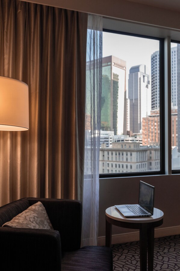 Habitación de hotel con vista a la ciudad, un sofá con un cojín estampado, una mesa redonda con una computadora portátil y un amplio ventanal con cortinas.