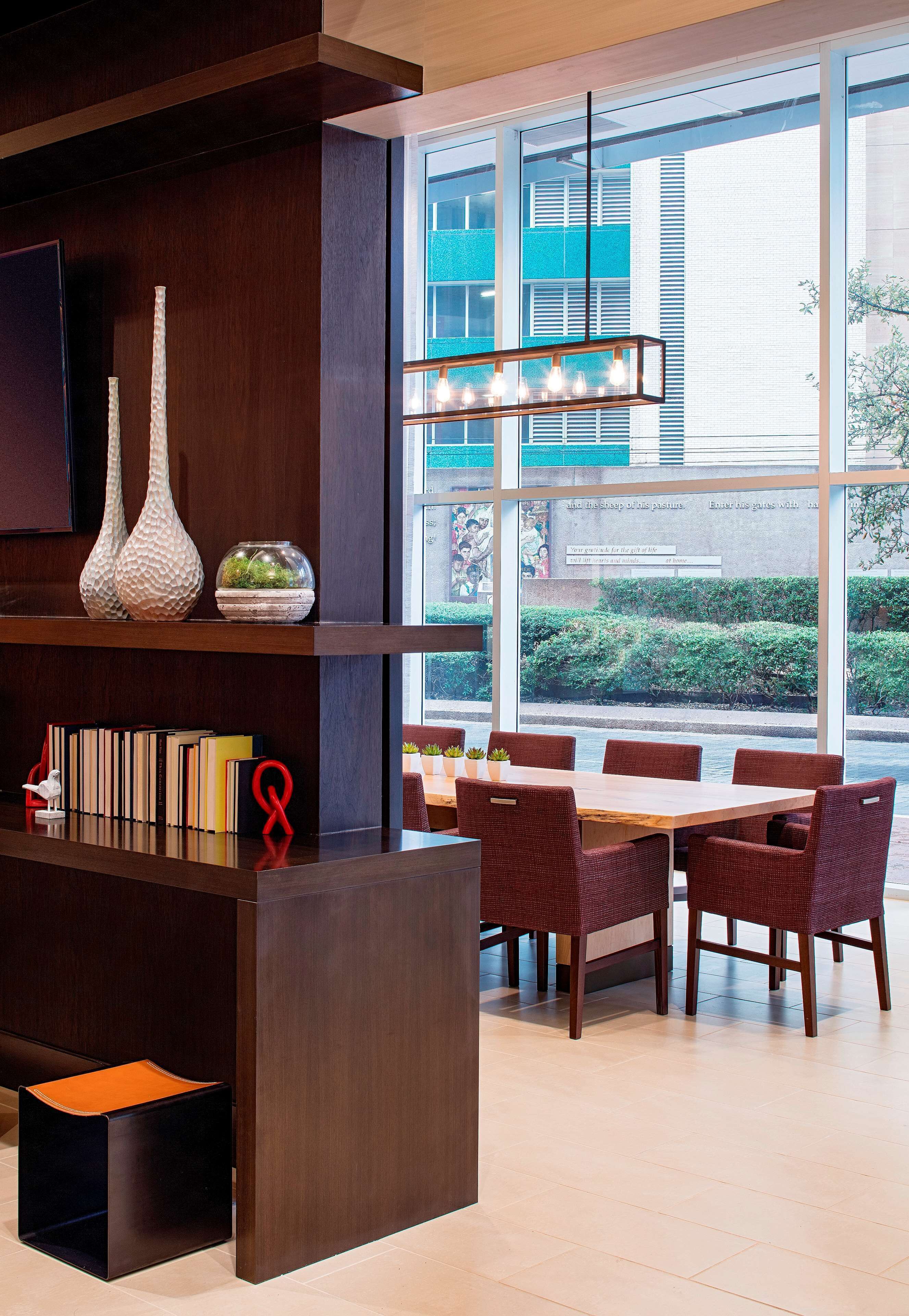 A modern dining area with a wooden table and wicker chairs, a bookshelf with books and decorative items, and a large window with a view of buildings and greenery.