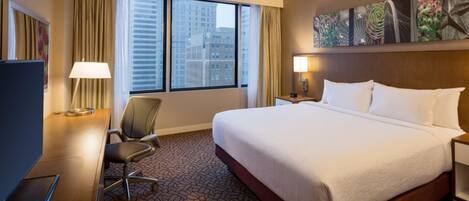 Una habitación de hotel con una cama grande, un escritorio con silla, una ventana con cortinas y una obra de arte colgada en la pared.