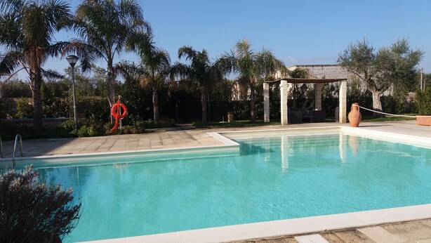 A clear swimming pool with a lifebuoy, a pergola, and palm trees. - Relais Tenuta Campì (Martano)