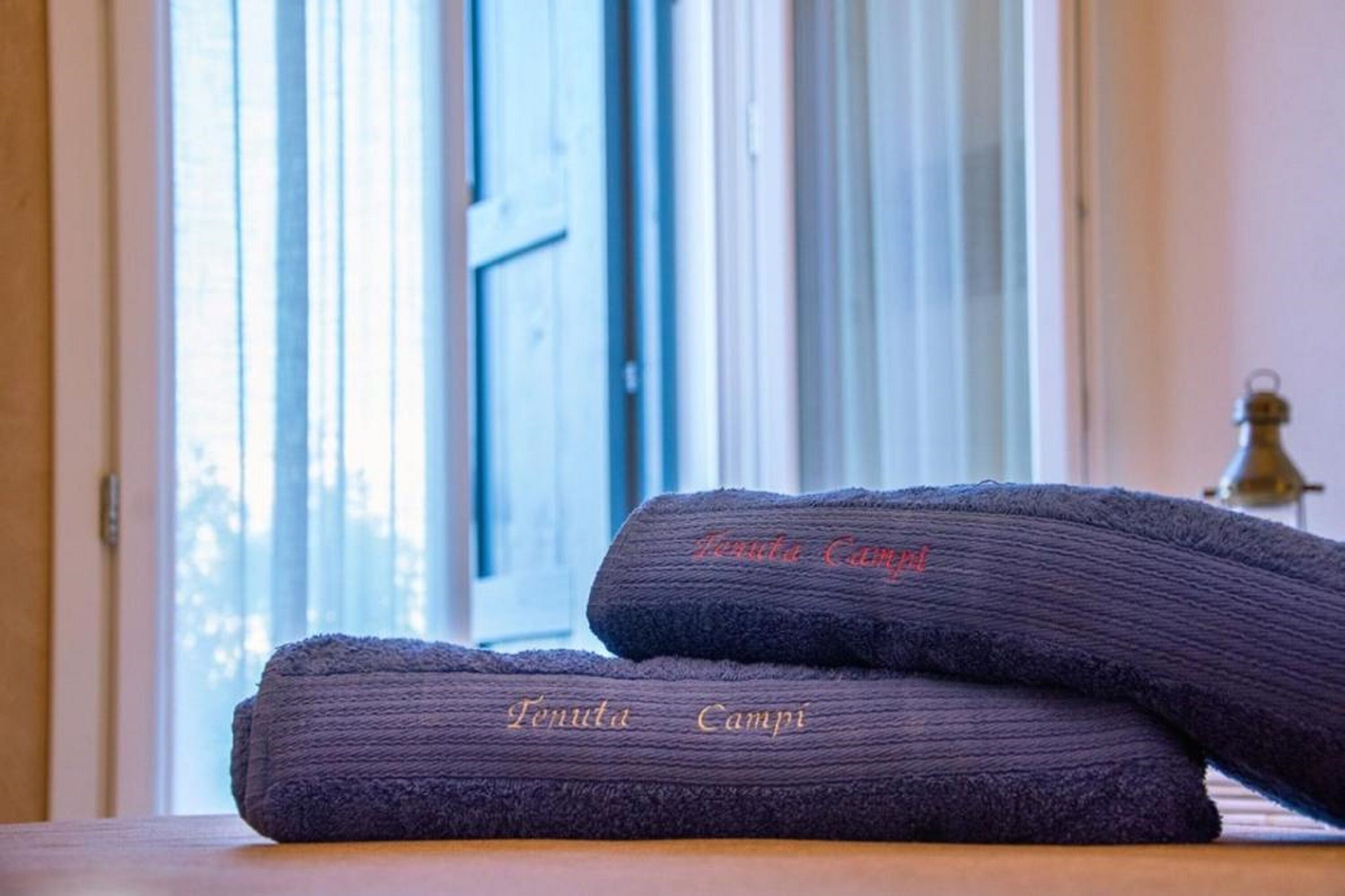 Two folded towels with "Fenuta Campi" embroidered on them, placed on a wooden surface near a window with a view of the outdoors.