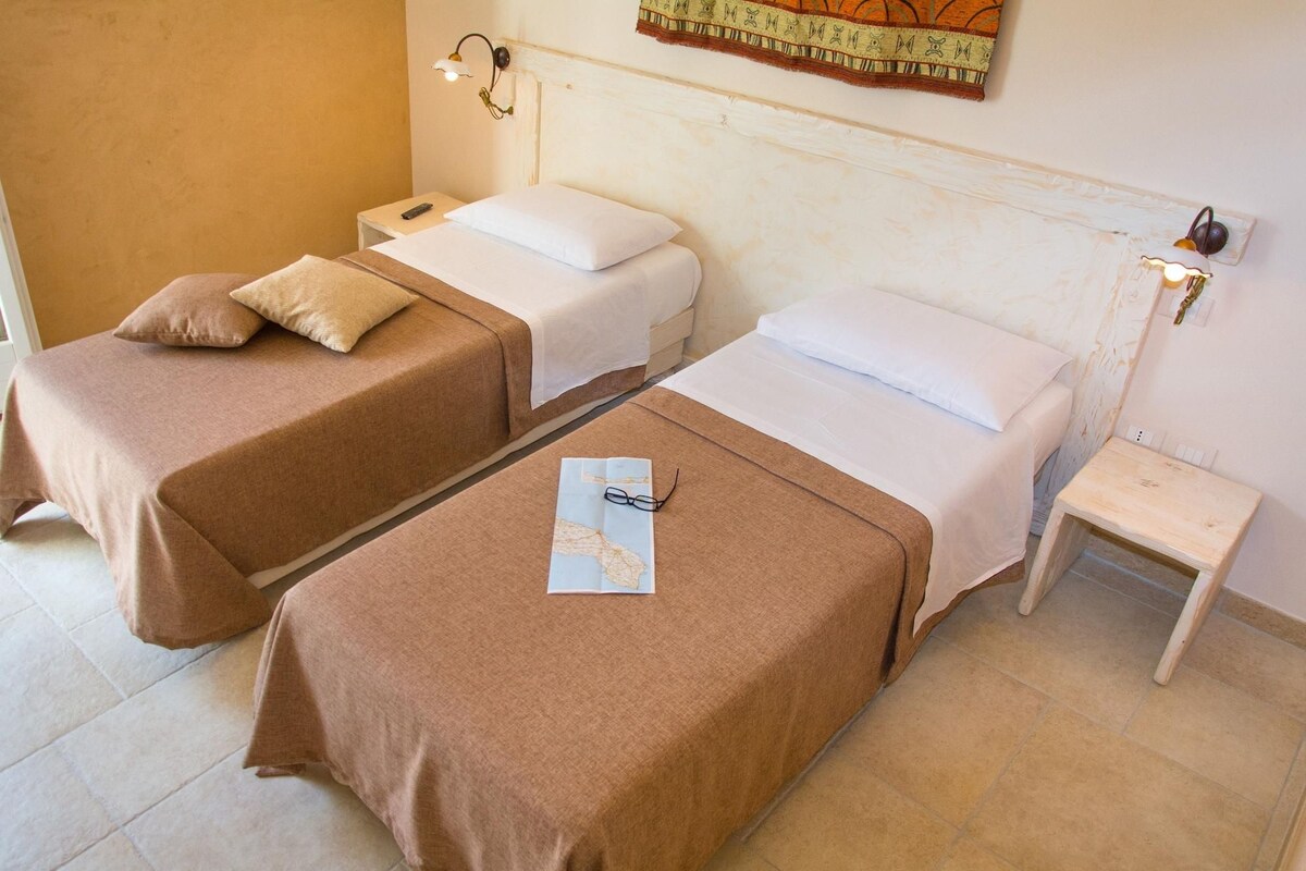 Una habitación con dos camas individuales, cada una con una colcha marrón y almohadas blancas. Hay una mesita de noche de madera con una lámpara y un control remoto encima. Un mapa y unos anteojos están sobre el suelo entre las camas.