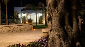 A well-lit building with a stone facade, a paved entrance, and a landscaped garden with flowers and a tree. - Relais Tenuta Campì (Martano)
