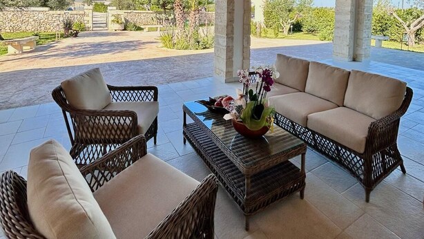 An outdoor patio with wicker furniture, a central table with a vase of flowers, and a view of a garden and buildings in the distance. - Relais Tenuta Campì (Martano)