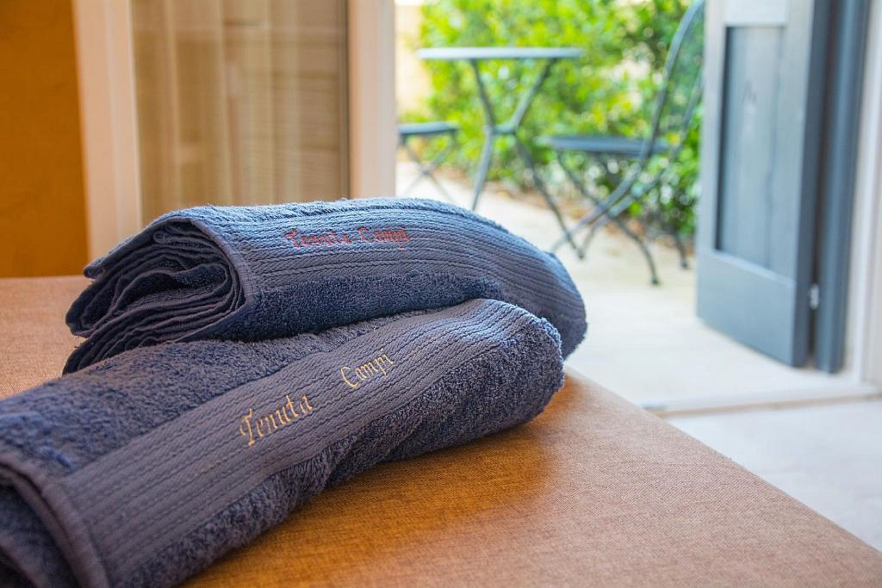 A stack of folded towels with embroidered text on a wooden surface, with a door and outdoor seating visible in the background.