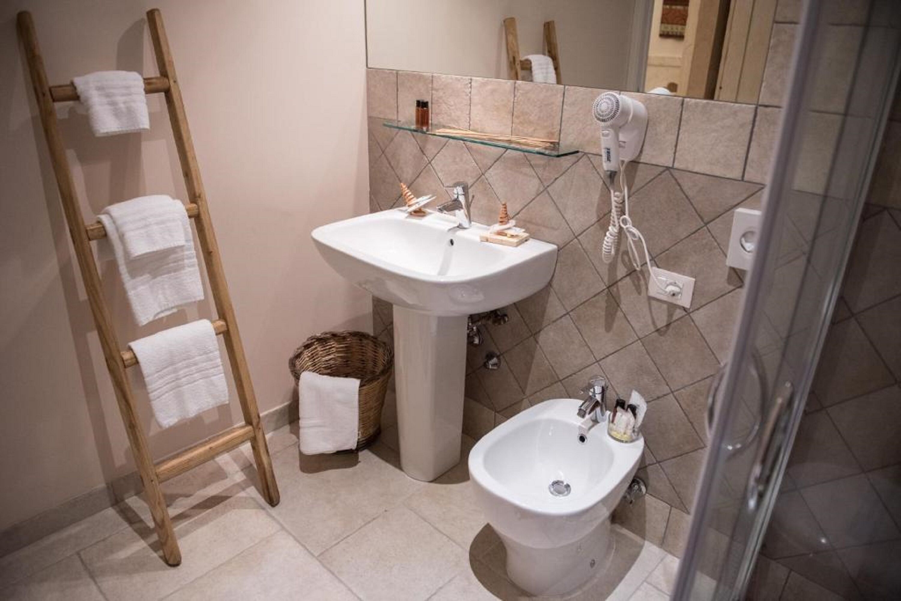 Un baño con un perchero de madera, un lavabo pedestal, un bidé y un estante de vidrio con artículos de aseo.