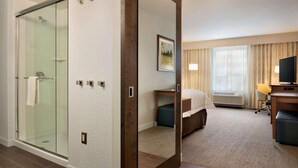 A hotel room with a glass shower enclosure, a bed with a headboard, a desk with a chair, and a television. - Hampton Inn Lincoln Airport (Lincoln)