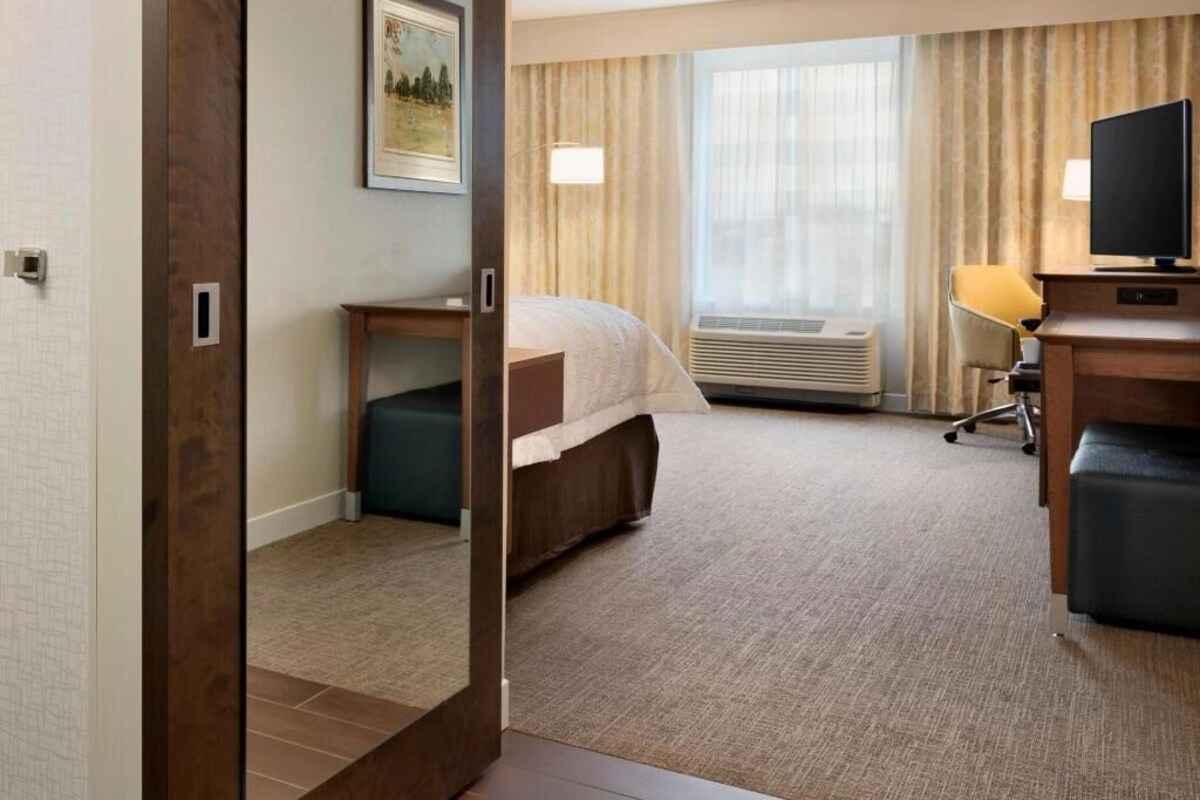 A hotel room with a bed, a desk, a chair, an air conditioning unit, and a window with curtains.