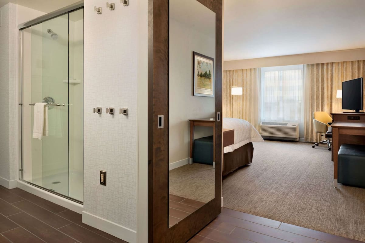 Habitación de hotel con cabina de ducha de vidrio, cama con cabecera, escritorio con silla y televisor.