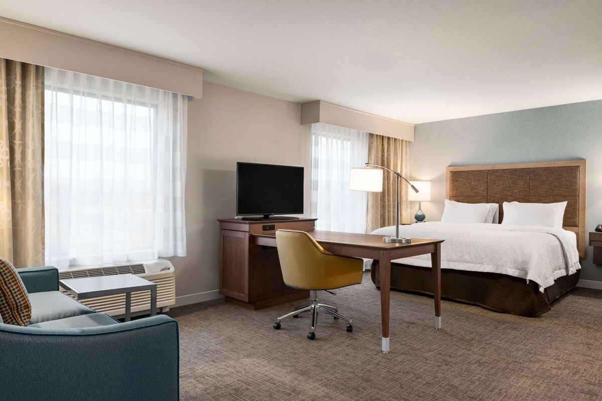 A hotel room with a bed, desk, chair, television, lamp, and large windows.