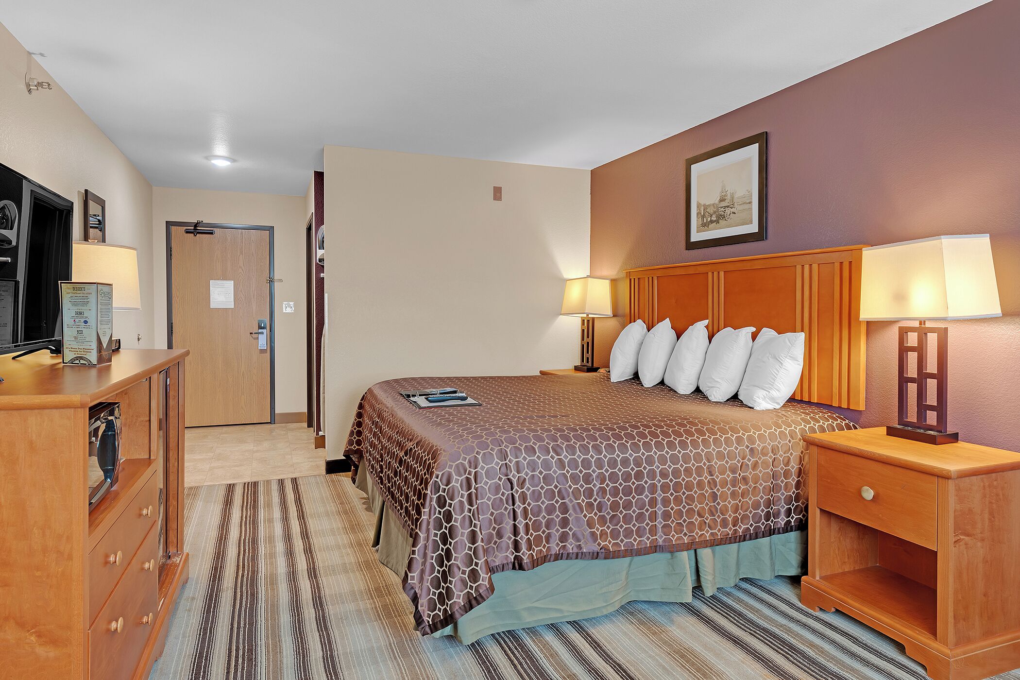 A hotel room with a bed, bedside tables, a dresser, a television, and a door.