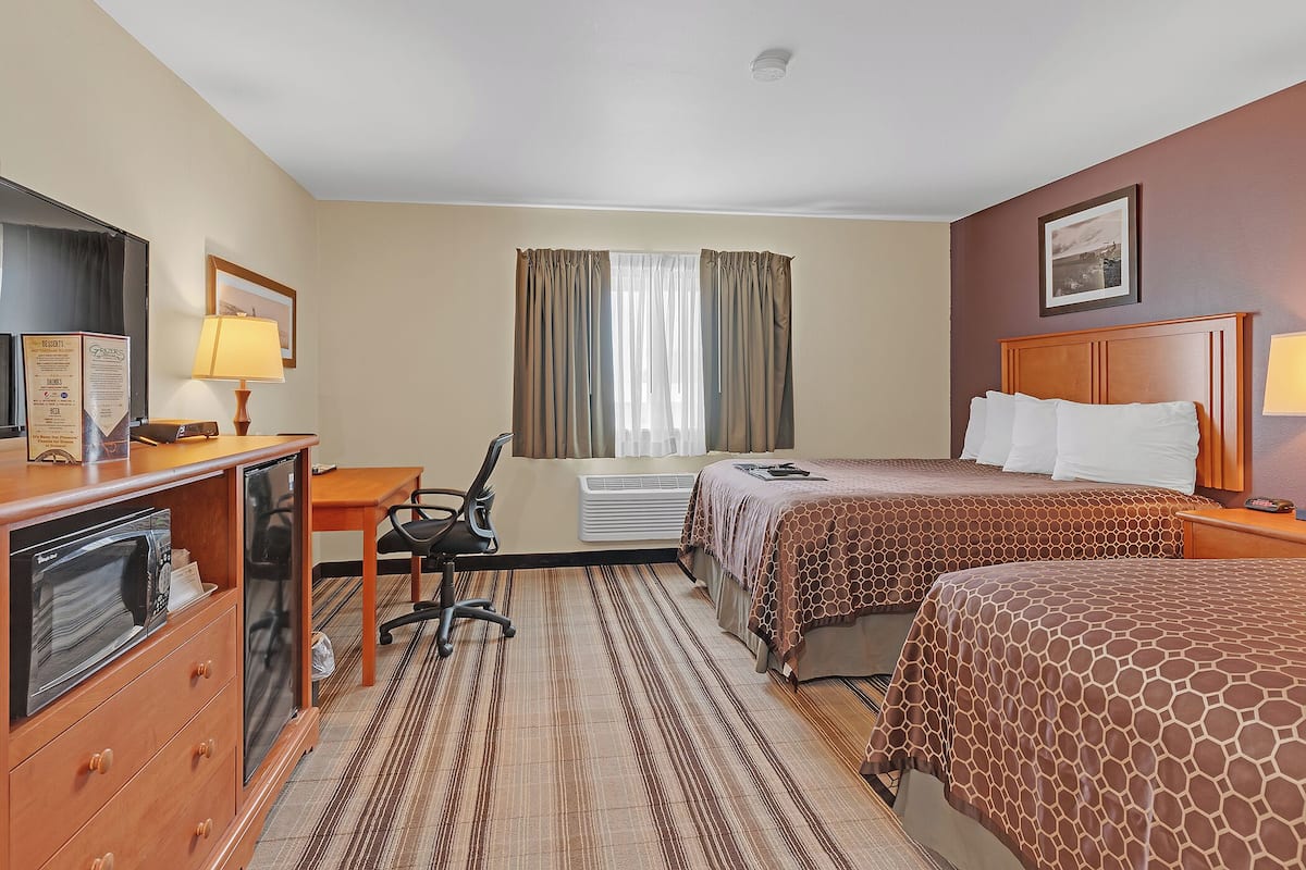 A hotel room with a desk, two beds, a chair, a microwave, a refrigerator, and a window with curtains.