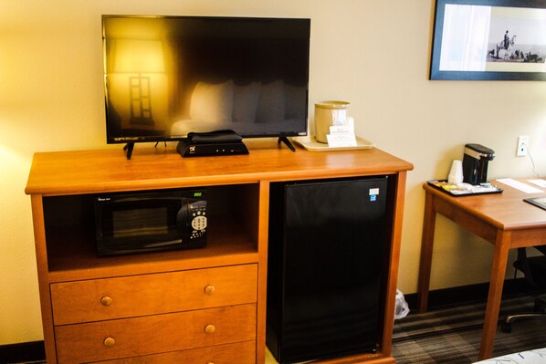 Habitación de hotel compacta con televisión de pantalla plana, microondas, refrigerador pequeño, cafetera y una mesita de madera.