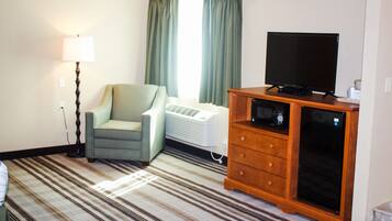 A hotel room with a striped carpet, a green armchair, a standing lamp, a flat-screen TV, a wooden dresser, and a white air conditioning unit.