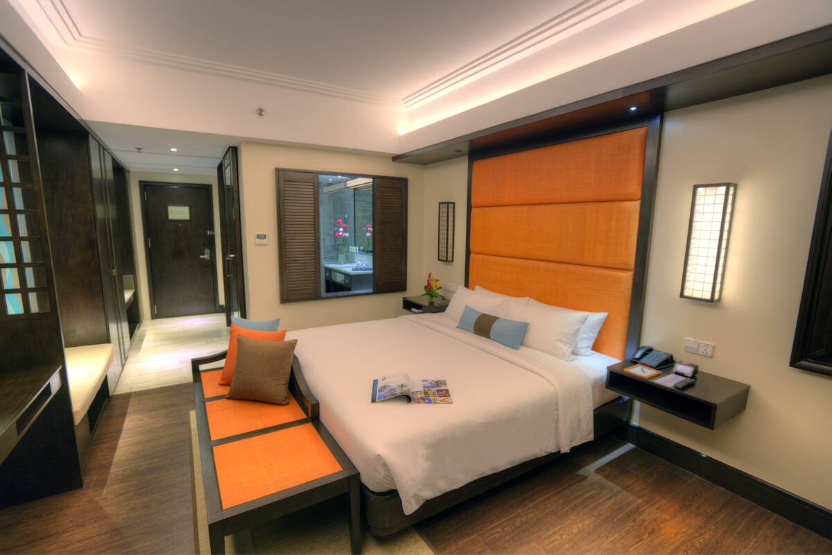 A modern hotel room with a large bed, a bedside table with a phone and a magazine, a bench with cushions, and a view of a bathroom through a mirror.