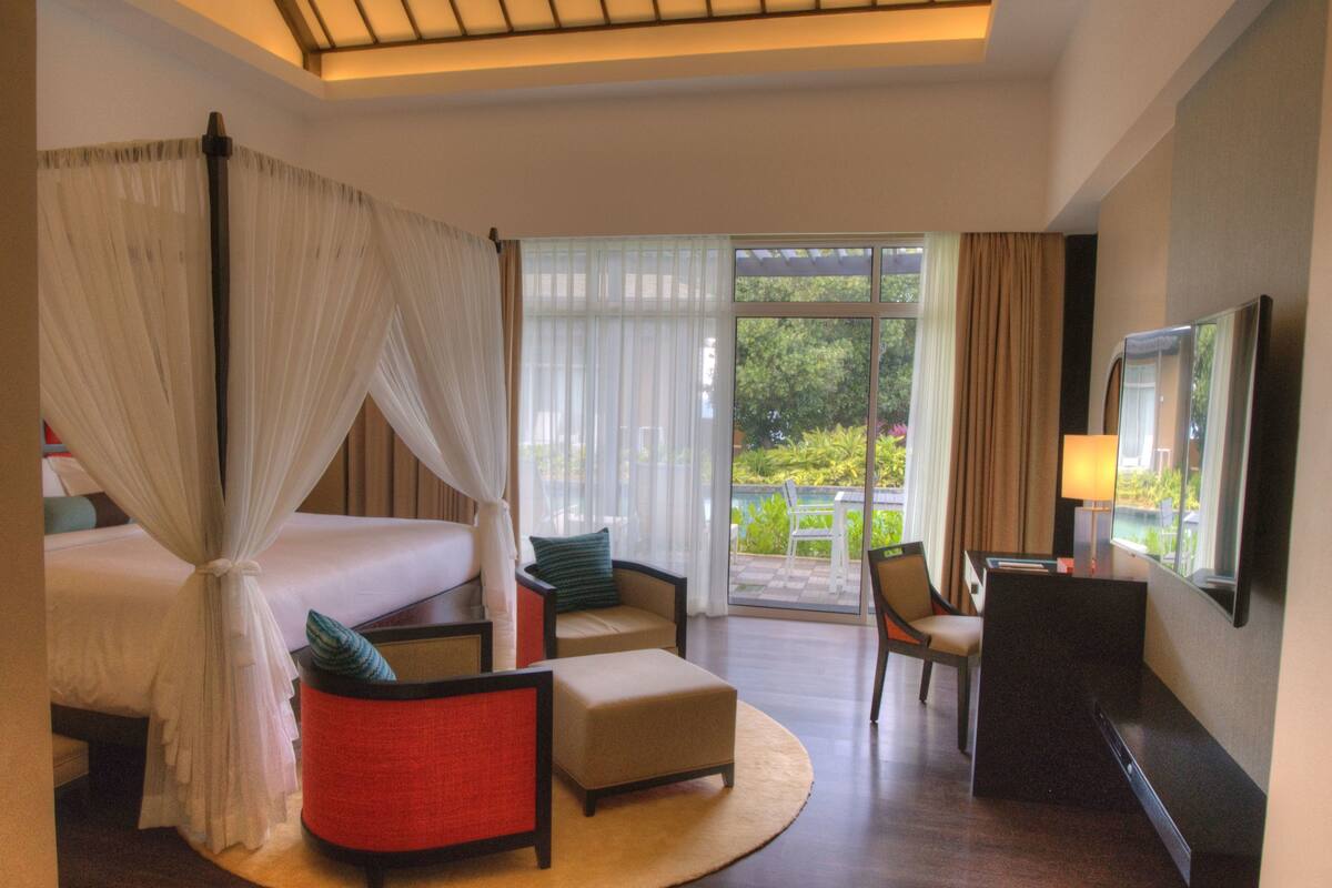 A hotel room with a bed, a sitting area with a chair and ottoman, a desk, and a large window with a view of greenery.
