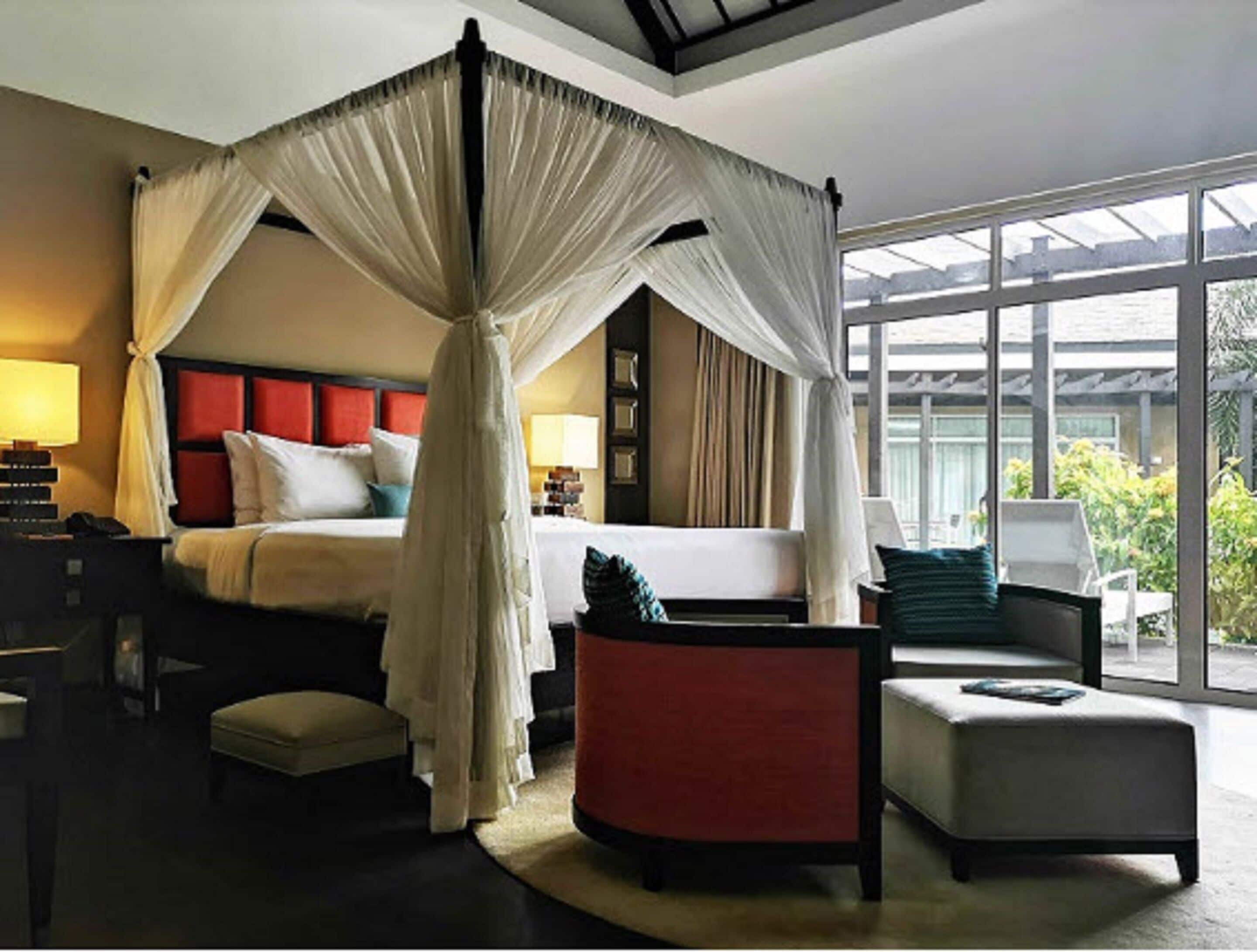 A four-poster canopy bed with a red headboard, a bedside table with a lamp, and a view of the outdoors through large windows.