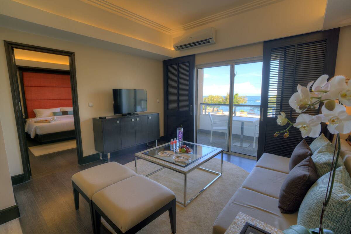 A hotel room with a bed, a TV, a glass coffee table with fruit, a sofa, and a view of the sea.