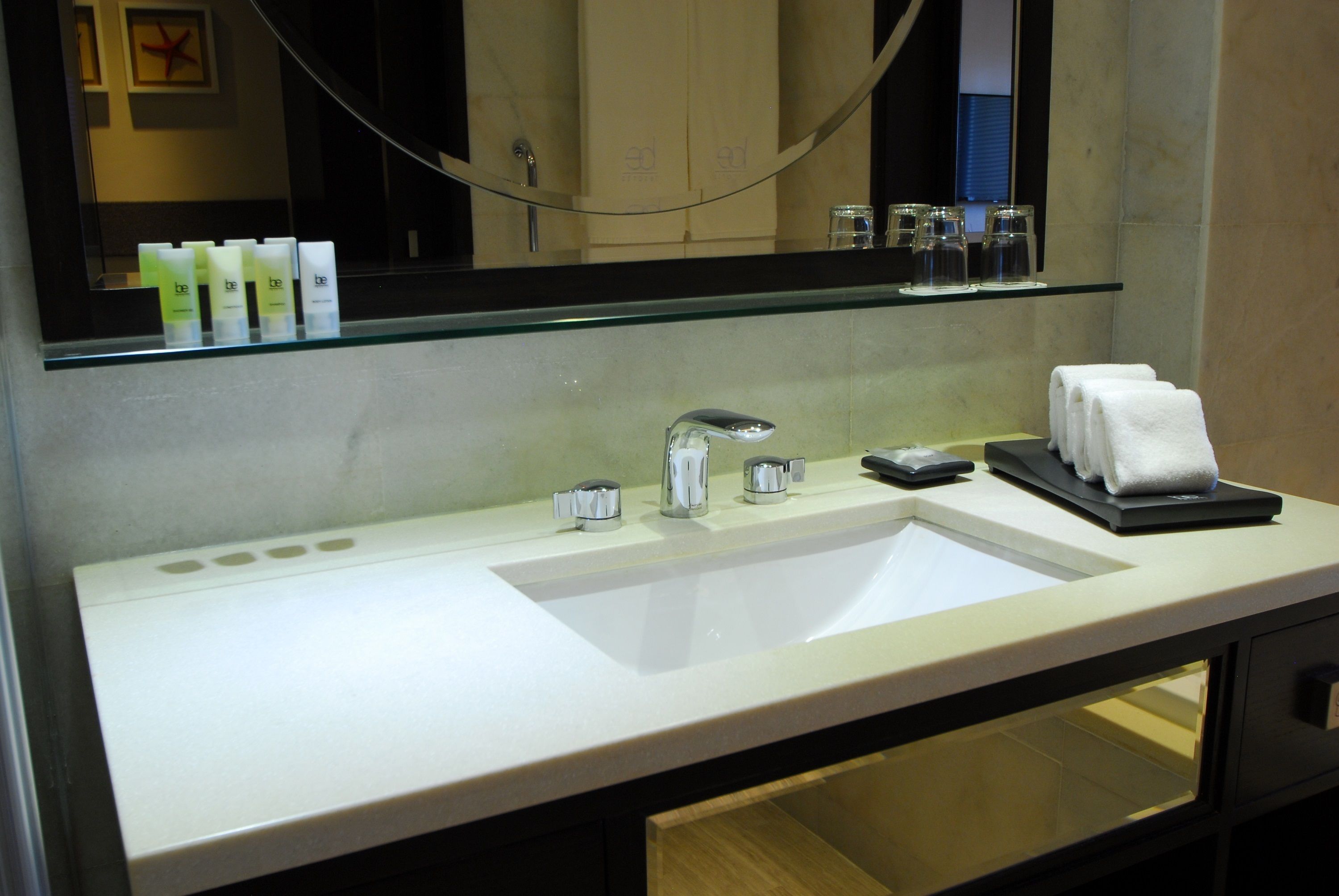 A hotel bathroom with a white sink, a mirror, and a glass shelf above the sink.