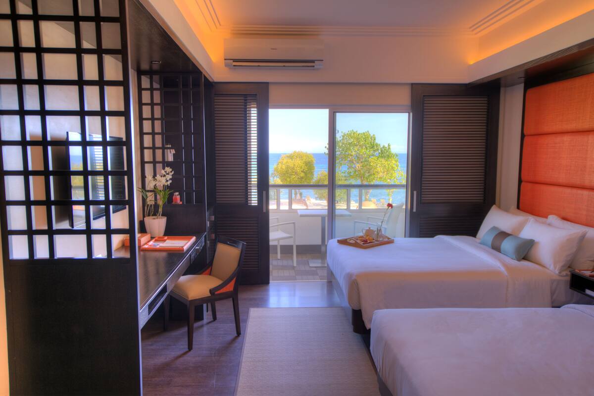 A hotel room with two beds, a desk with a chair, and a view of the sea.
