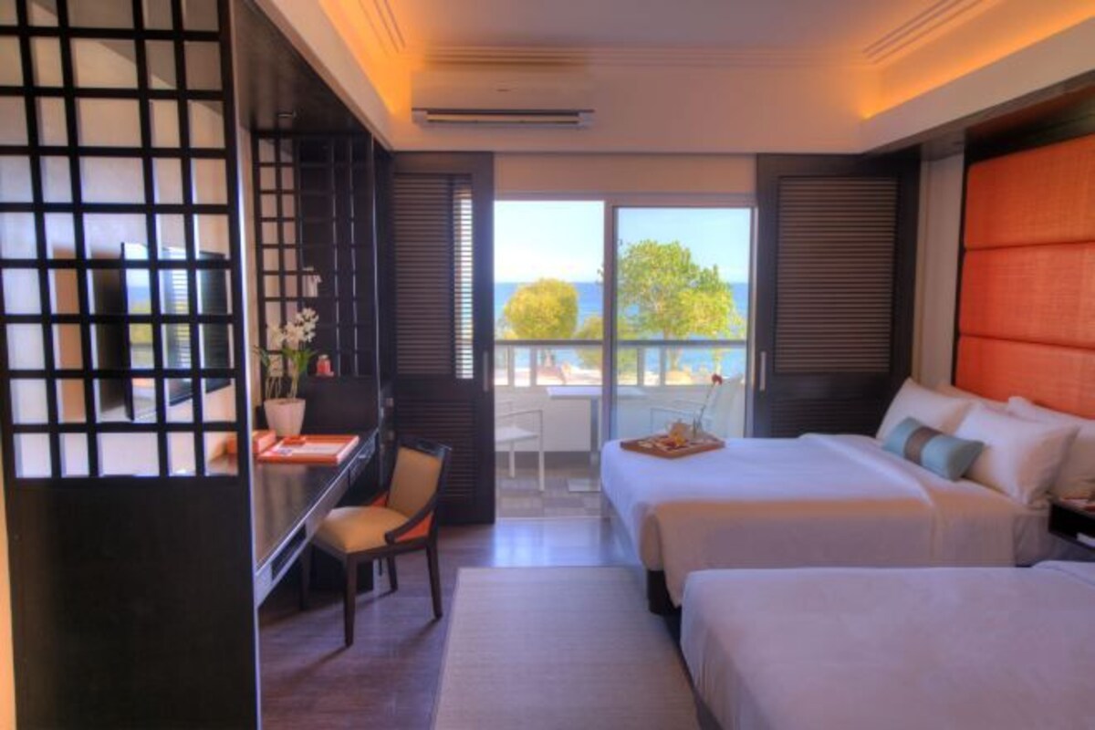 A hotel room with two beds, a desk with a chair, a view of the sea, and a balcony.
