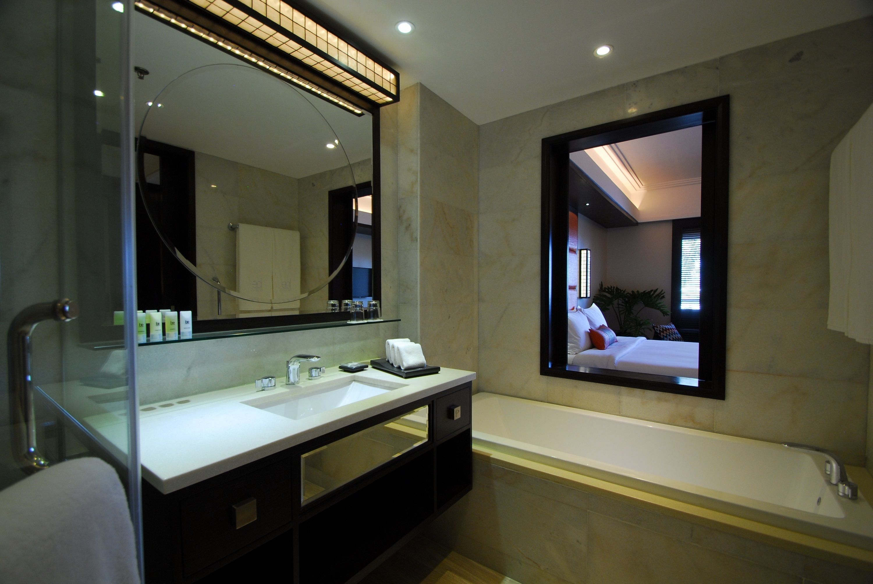 A modern bathroom with a large mirror, a white sink, a bathtub, and a television screen displaying a bedroom.
