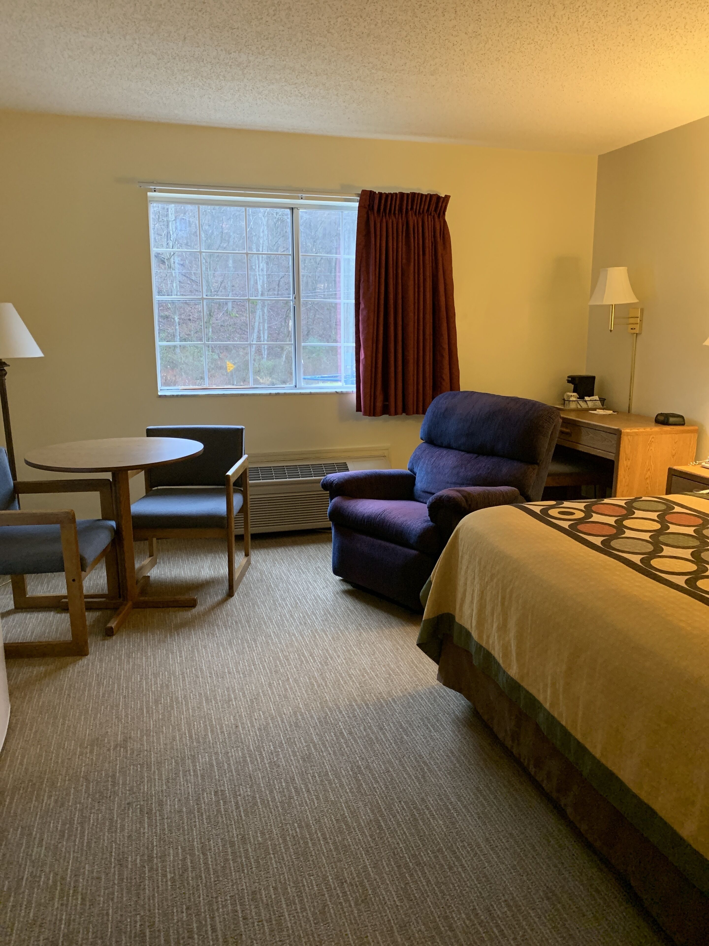 A hotel room with a bed, a desk with a chair, a purple armchair, a window with curtains, and a lamp.