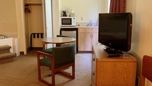 Ett kompakt hotellrum med ett litet pentry, en matplats med bord och stolar, en tv på ett träställ och en röd stol.