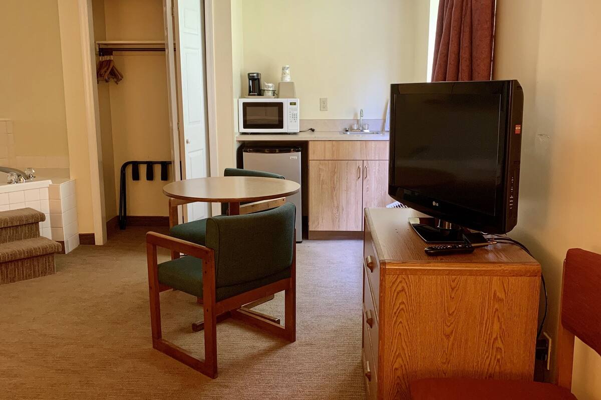 Quarto de hotel compacto com cozinha compacta, área de refeições com mesa e cadeiras, televisão sobre um suporte de madeira e uma cadeira vermelha.
