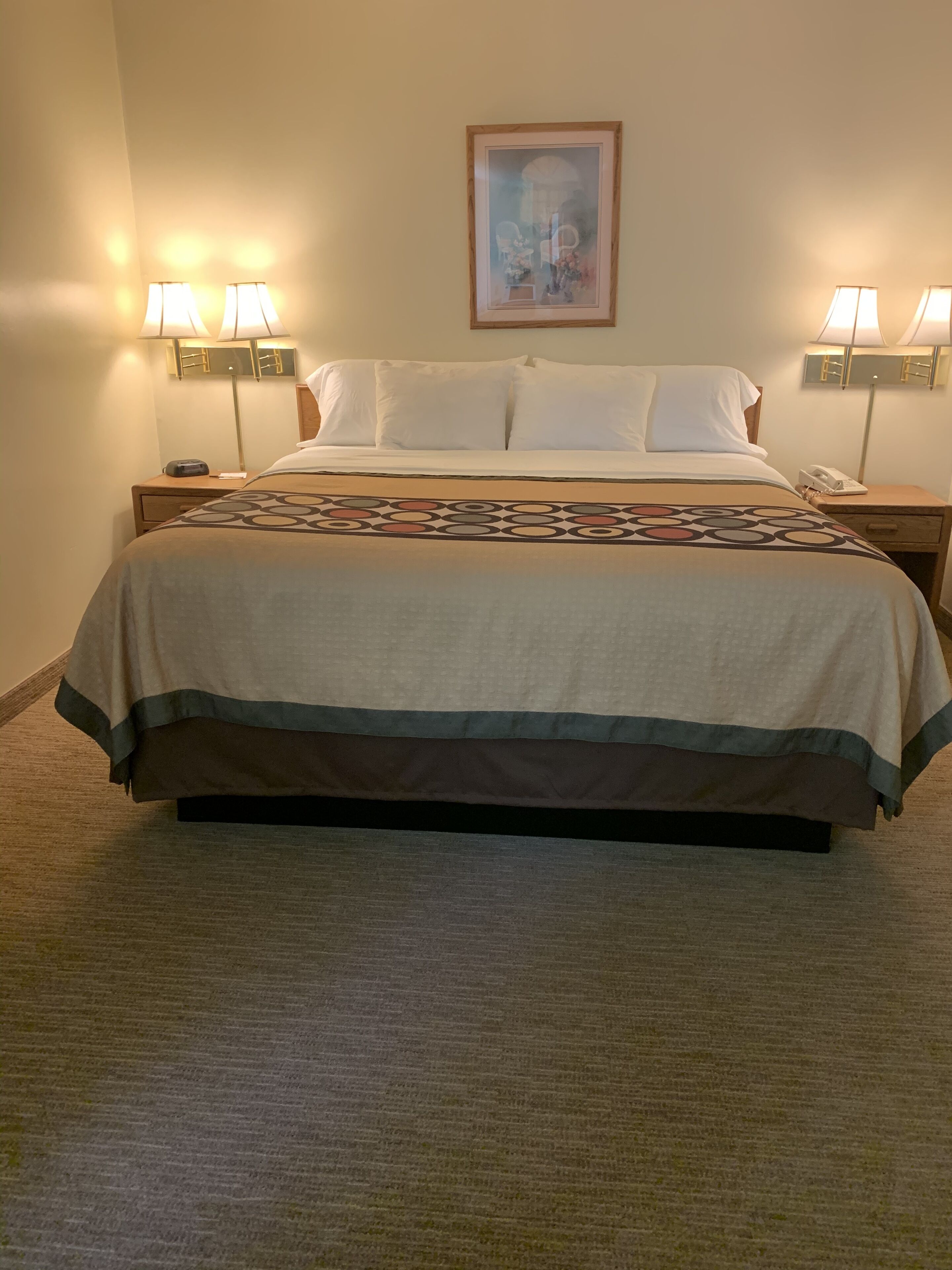 A hotel room with a large bed, two bedside tables with lamps, a framed picture on the wall, and a carpeted floor.