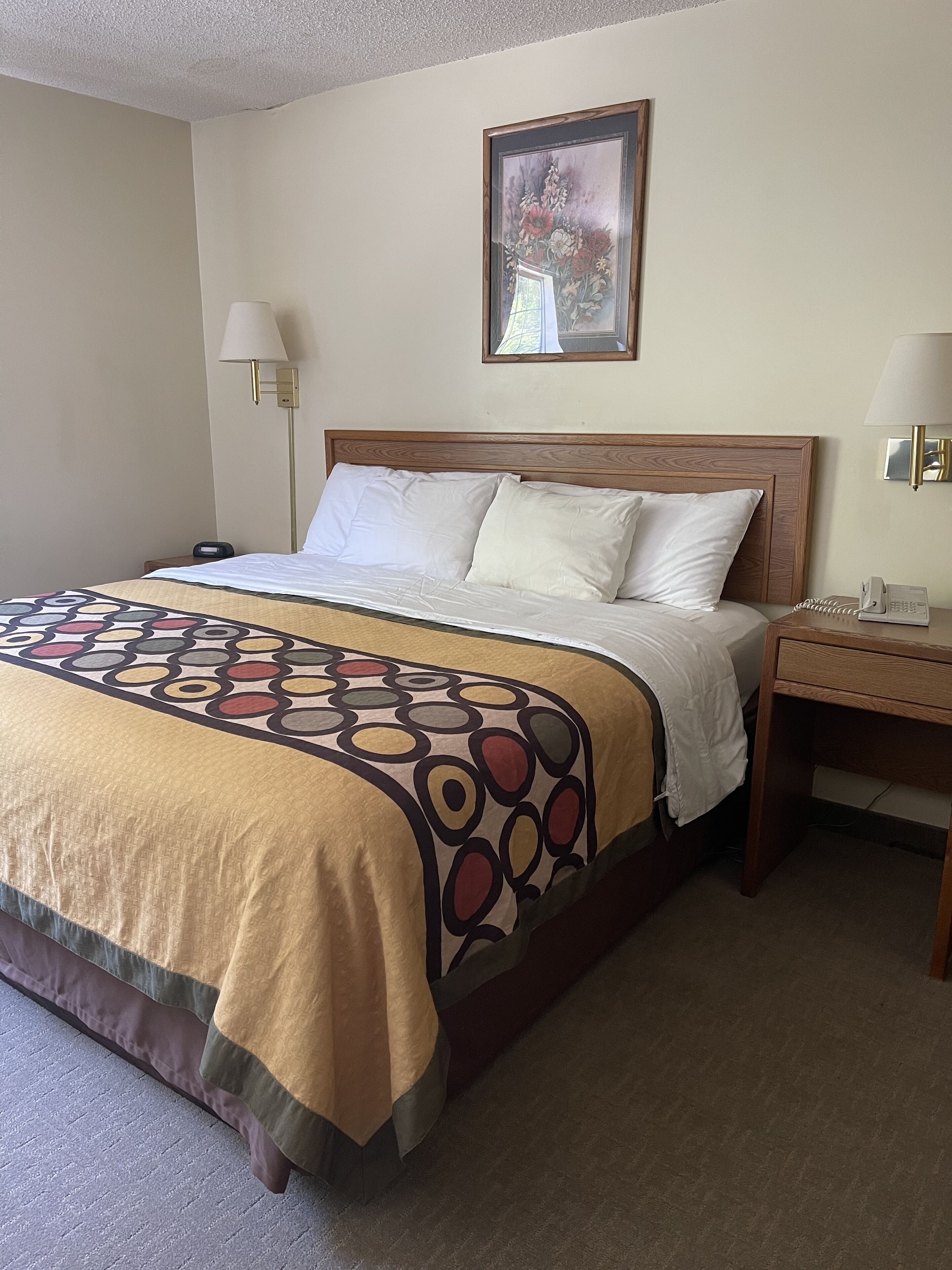 A hotel room with a wooden headboard, a large bed with white linens, a patterned throw blanket, a bedside table with a lamp, and a framed picture on the wall.