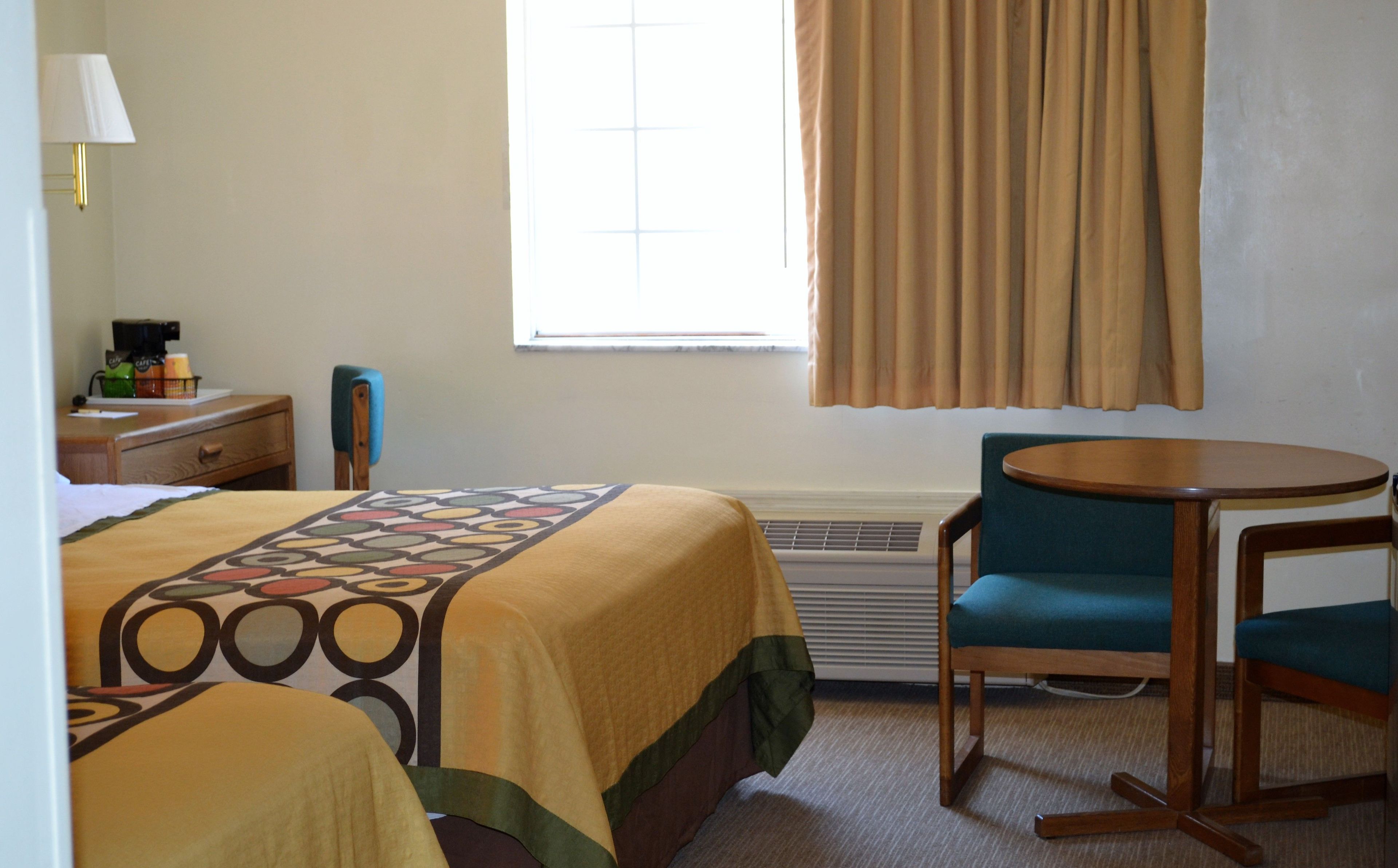 A hotel room with a bed, a desk with a lamp, a chair, a small table, and a window with curtains.