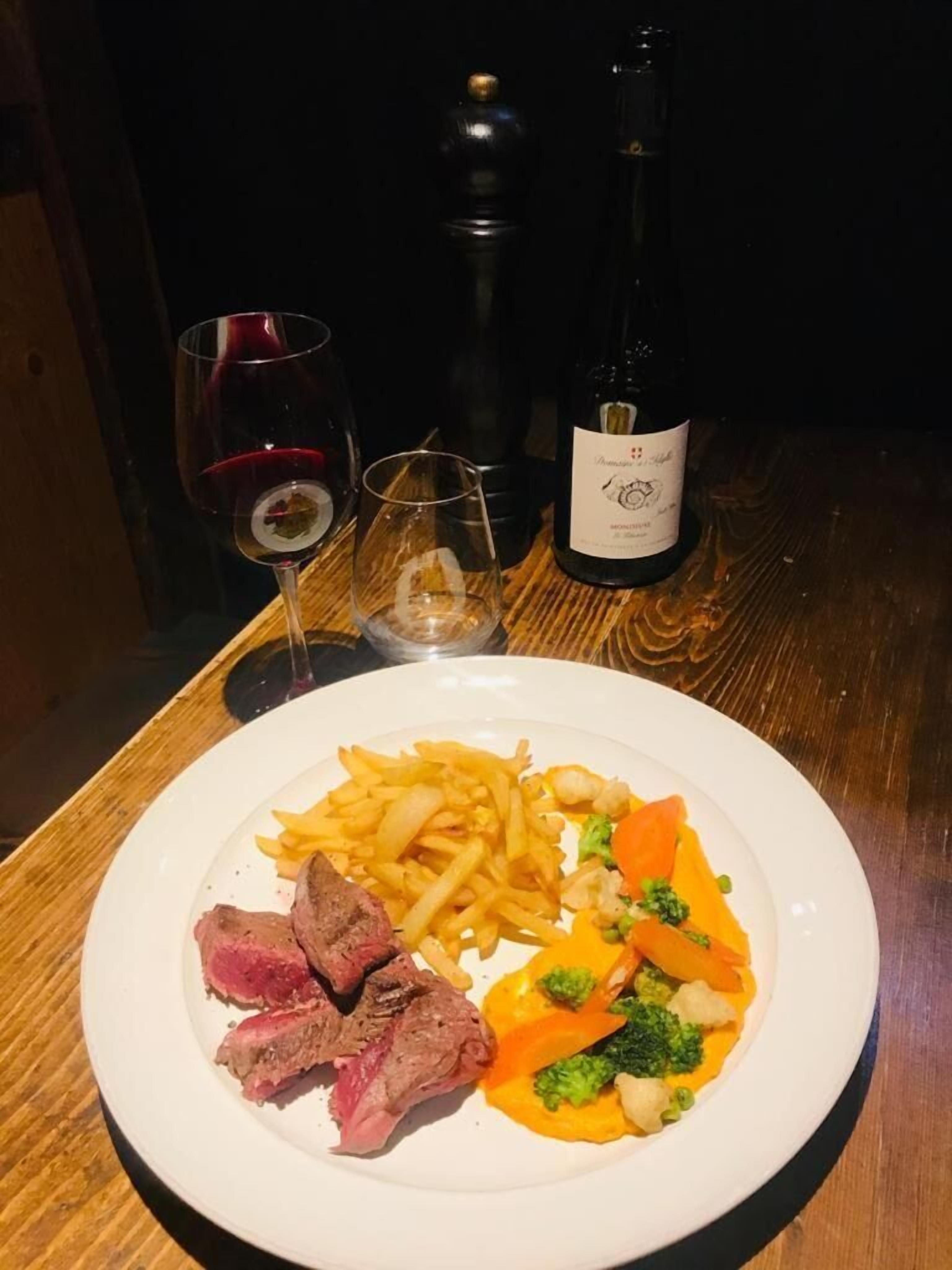 Un plato de comida con carne, papas fritas y vegetales, una botella de vino y dos copas sobre una mesa de madera.