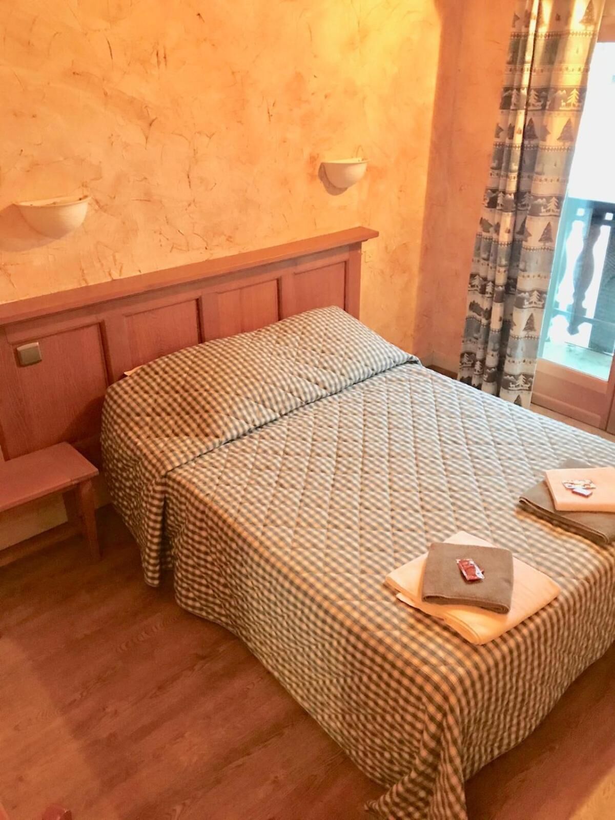 A bedroom with a checkered bedspread, a bedside table, a nightstand, and a window with curtains.