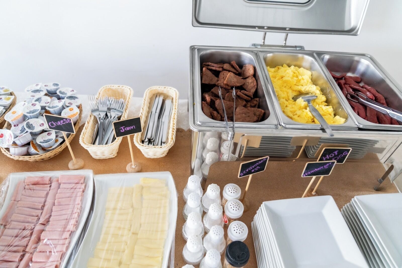 A buffet with labeled sections for cheese, jam, scrambled eggs, hot dogs, and ham.