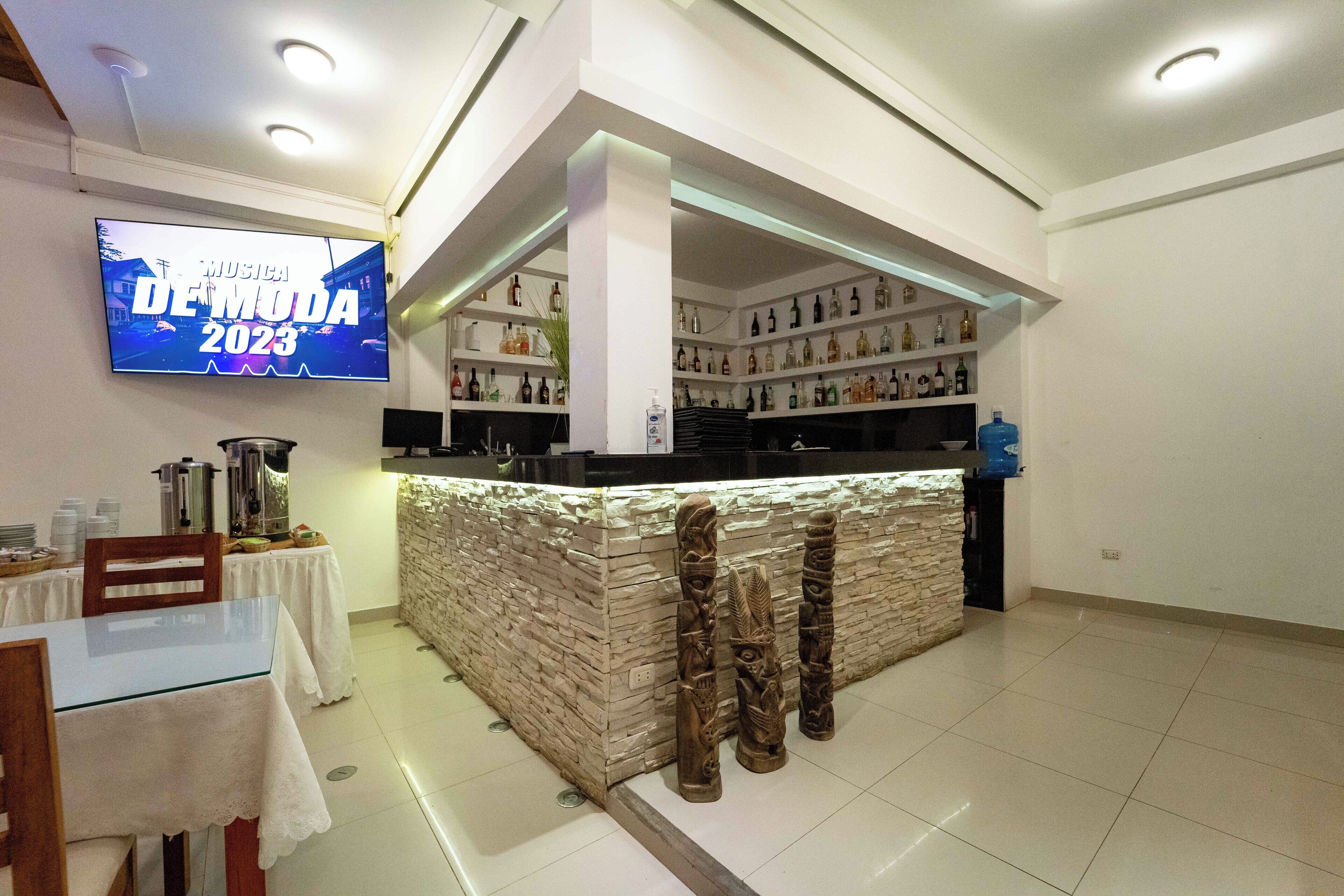Zona de bar con mostrador de piedra, estanterías con botellas y un televisor mostrando 'MUSICA DE MODA 2023'.